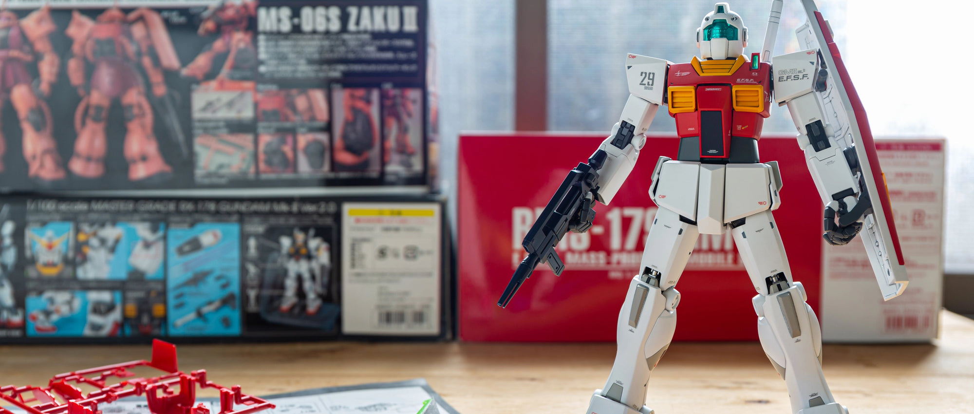 A assembled white, red, and blue Gundam model figure stands on a wooden table with its original boxes in the background, alongside unassembled red plastic parts, tools, and a manual in the foreground.